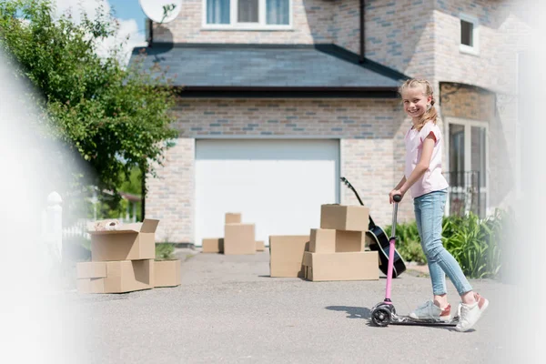 Niño sonriente cabalgando en patinete scooter cerca de cajas y guitarra acústica en frente de la nueva casa - foto de stock