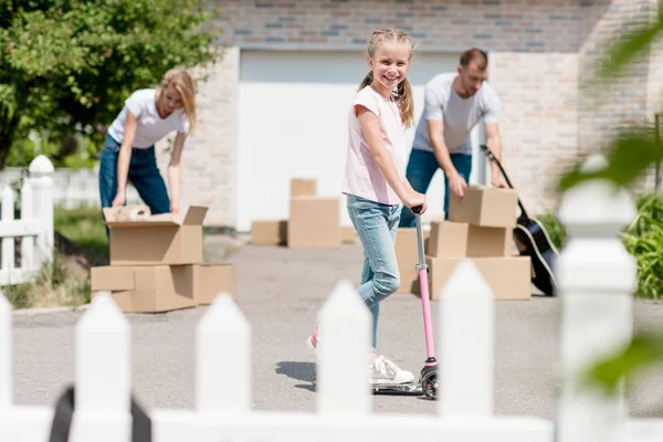 Pareja desempacar cajas de cartón mientras su hija sonriente montar en patinete scooter delante de la nueva casa de campo - foto de stock