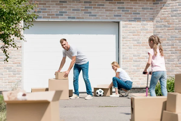 Young couple unpacking cardboard boxes while their daughter riding on kick scooter in front of new cottage — Stock Photo