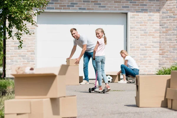 Pareja desempacar cajas de cartón mientras que su hija montar en patada scooter delante de la nueva casa de campo - foto de stock