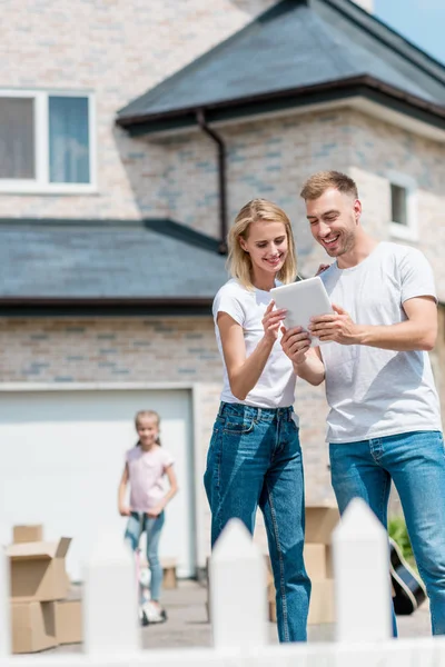 Smiling couple using digital tablet while their daughter riding on kick scooter behind in yard of new house — Stock Photo