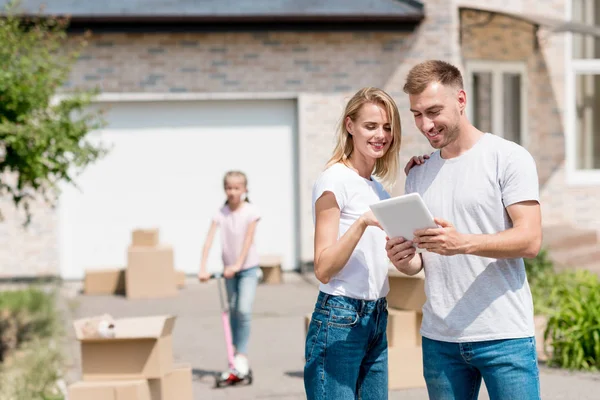 Feliz pareja usando digital tablet mientras su hija cabalgando en kick scooter detrás en patio de nueva casa — Stock Photo