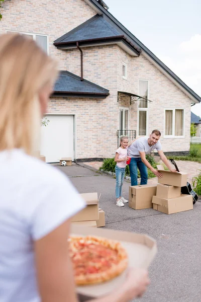 Image recadrée d'une femme tenant une pizza tandis que son mari et sa fille déballent des boîtes en carton devant une nouvelle maison — Photo de stock