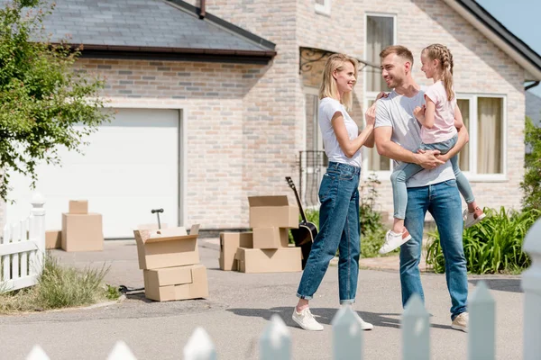 Happy woman embracing smiling husband while he holding daughter inf front of new house — Stock Photo