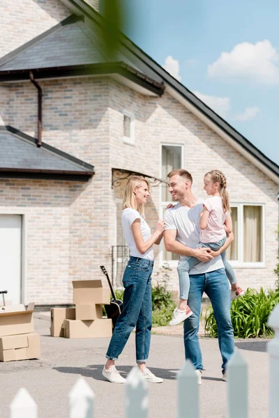 Attractive woman embracing smiling husband while he holding daughter inf front of new house — Stock Photo