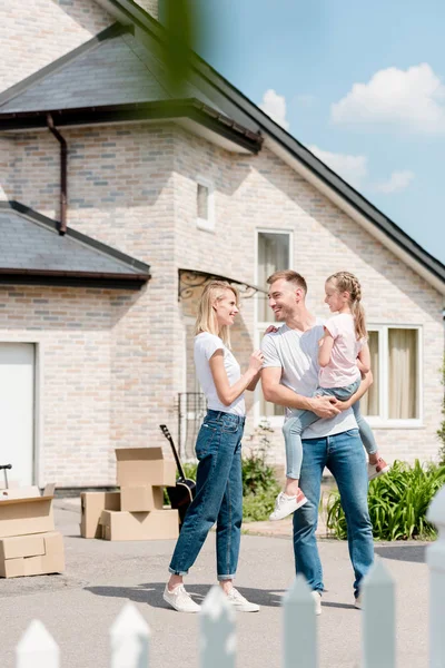 Atractiva mujer abrazando sonriente marido mientras él sosteniendo hija inf frente a nueva casa — Stock Photo