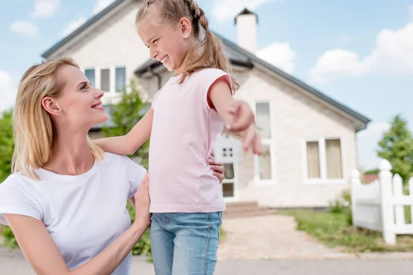 Nahaufnahme des Schlüssels mit Schmuckstück in der Hand des lächelnden kleinen Kindes, das die Mutter vor dem neuen Haus umarmt — Stockfoto