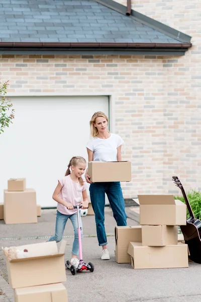 Mother unpacking cardboard boxes and little daughter riding on kick scooter in front of new cottage — Stock Photo