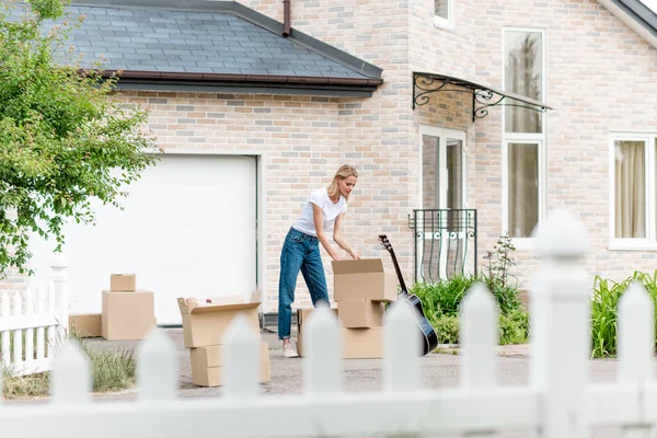Femme déballage boîtes en carton près de la guitare devant une nouvelle maison — Photo de stock