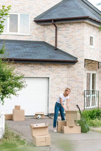 Jovem mulher desempacotando caixas de papelão perto de guitarra na frente de nova casa de campo — Fotografia de Stock