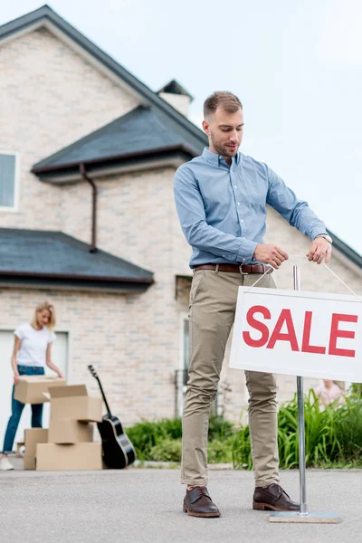 Corretor de imóveis masculino pendurado sinal de venda na frente da mulher embalagem de caixas de papelão para se mudar de casa — Fotografia de Stock