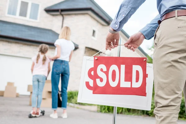 Vue partielle de l'agent immobilier suspendu panneau vendu devant les gens qui déménagent dans une nouvelle maison — Photo de stock