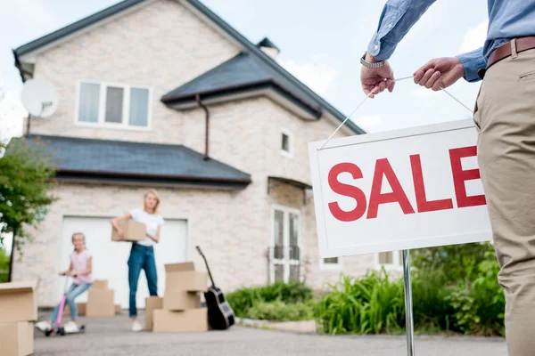 Imagen recortada de agente inmobiliario masculino con letrero de venta y familia con cajas de cartón detrás - foto de stock