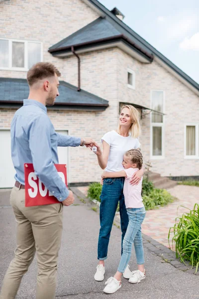 Sorridente agente immobiliare maschile con segno venduto dando la chiave alla giovane donna con figlia di fronte alla nuova casa — Foto stock