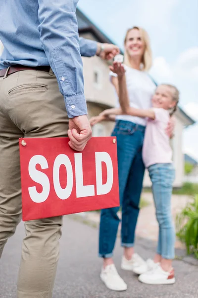 Imagen recortada de agente inmobiliario con letrero vendido que da la llave a la mujer joven con hija en frente de la nueva casa - foto de stock