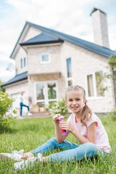 Очаровательный маленький ребенок с мыльными пузырьками, сидящий на газоне, в то время как ее мать стоит позади перед домом — стоковое фото