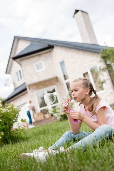 Vue latérale du petit enfant avec des bulles de savon assis sur la pelouse tandis que sa mère se tient derrière devant la maison — Photo de stock