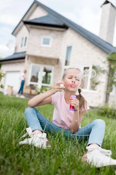 Маленькая девочка использует пузырьковый воздуходувку и сидит на газоне, в то время как ее мать стоит позади дома — стоковое фото