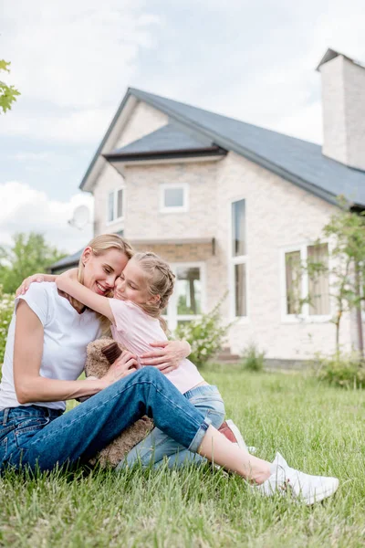 Pequeña hija abrazando sonriente madre en el césped verde en frente de la casa de campo - foto de stock