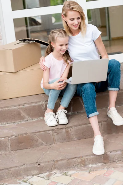 Donna felice con figlia che usa il computer portatile sulle scale con scatole di cartone e chitarra di fronte alla nuova casa — Foto stock