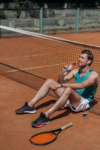 Jeune homme heureux assis sur le sol et boire de l'eau après la formation de tennis — Photo de stock