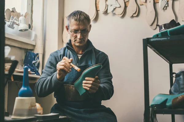 Zapatero maduro sosteniendo la pieza de trabajo de la bota y trabajando con suela en su taller tradicional - foto de stock