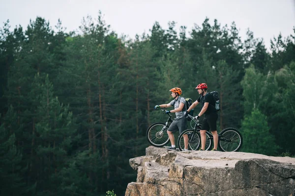Vista lateral de los jóvenes ciclistas de trial de pie sobre un acantilado rocoso con un bosque de pinos borroso en el fondo y apuntando a algún lugar - foto de stock