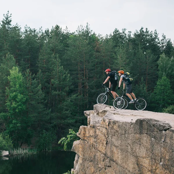 Vista lateral de los jóvenes ciclistas de trial balanceándose sobre un acantilado rocoso con un bosque de pinos borroso en el fondo - foto de stock