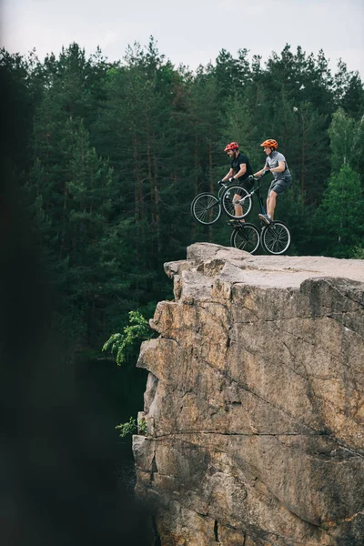 Vue latérale des motards d'essai debout sur des roues arrière sur une falaise rocheuse avec forêt de pins floue en arrière-plan — Photo de stock