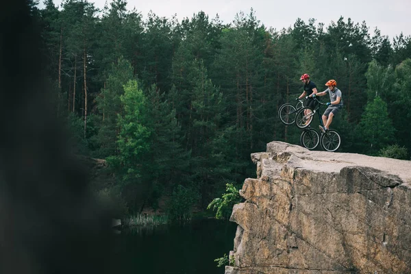 Side view of extreme trial bikers standing on back wheels on rocky cliff with blurred pine forest on background — Stock Photo