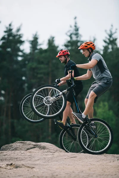 Handsome young trial bikers standing on back wheels on rocky cliff with blurred pine forest on background — Stock Photo