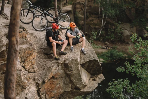 Vista de ángulo alto de jóvenes ciclistas de trial activos relajándose en acantilado rocoso después del paseo - foto de stock