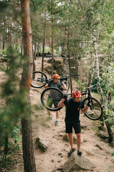 Young trial bikers carrying bikes on backs at forest — Stock Photo