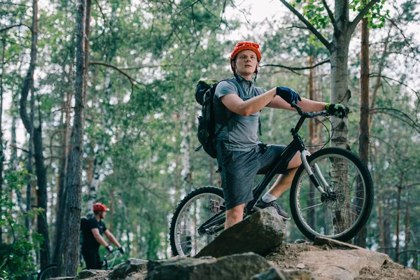 Young trial biker standing on rock at beautiful forest and looking away with his friend blurred on background — Stock Photo