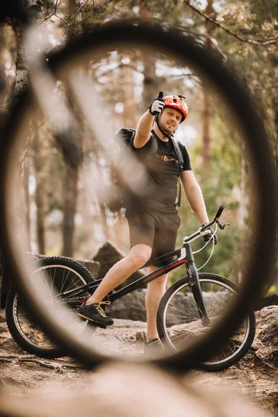 View of young trial biker showing thumb up at forest through bike wheel — Stock Photo