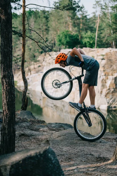 Young trial biker balancing on back wheel outdoors in front of lake at forest — Stock Photo