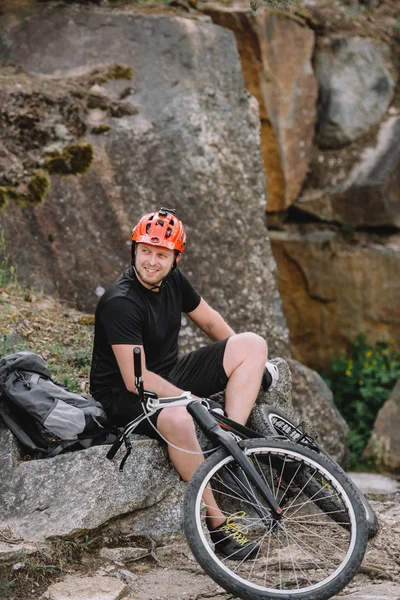 Happy young trial biker relaxing on rocks with bicycle outdoors — Stock Photo