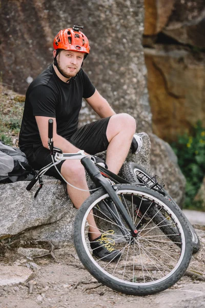 Handsome young trial biker relaxing on rocks with bicycle outdoors and looking at camera — Stock Photo