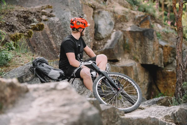 Joven ciclista de trial relajándose en las rocas con bicicleta al aire libre y mirando hacia otro lado - foto de stock