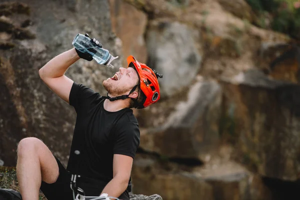 Handsome young traveller in helmet drinking water from plastic bottle in front of rocks — Stock Photo