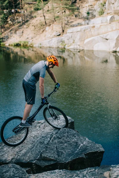 Athletic trial biker balancing on rocky cliff over lake — Stock Photo