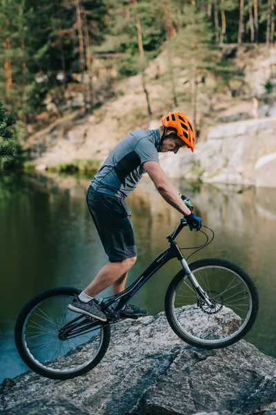 Young trial biker balancing on rocky cliff over lake — Stock Photo