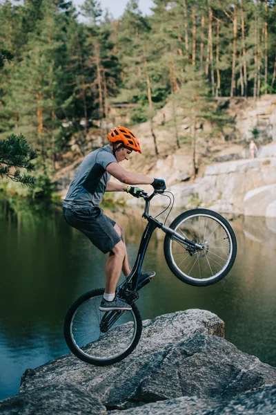 Athletic trial biker balancing on back wheel on rocky cliff over lake — Stock Photo