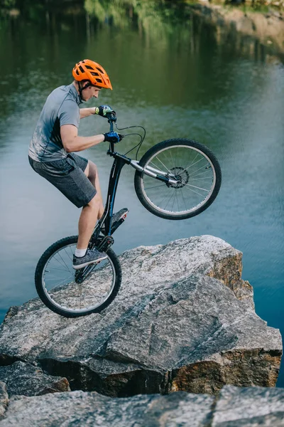 Ciclista de prueba extrema equilibrio en la rueda trasera en acantilado rocoso sobre el lago - foto de stock