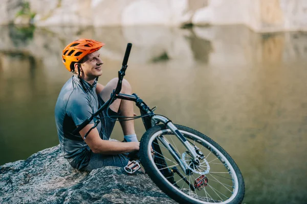 Smiling young trial biker relaxing on rocky cliff over water — Stock Photo