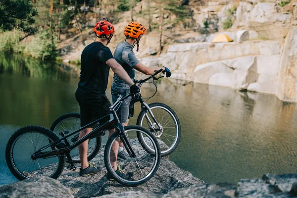 Extreme trial bikers standing on rocky cliff in front of mountain lake — Stock Photo