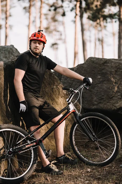 Handsome young trial biker leaning back on rocks outdoors in forest and looking at camera — Stock Photo