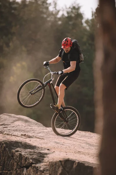 Handsome trial biker balancing on back wheel on rocks outdoors — Stock Photo