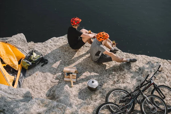 Vista de ángulo alto de los viajeros en bicicleta activa comer comida enlatada en acantilado rocoso sobre el lago - foto de stock