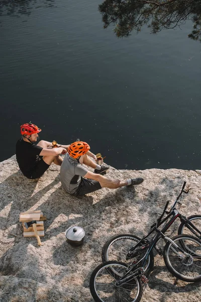 Vista de ángulo alto de los jóvenes ciclistas de trial comiendo comida enlatada en un acantilado rocoso - foto de stock